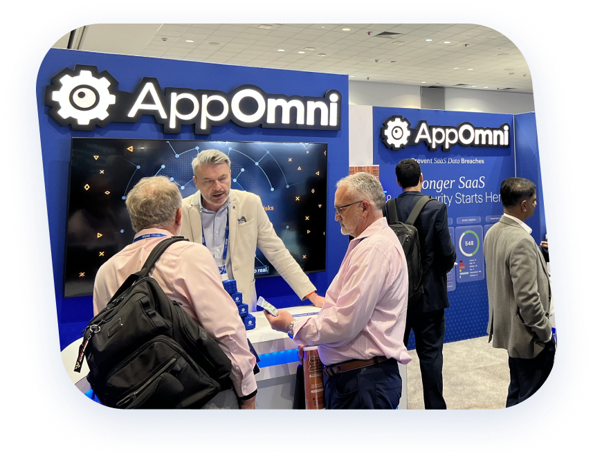 A group of men engage in conversation at the AppOmni booth during a tech event. The booth features a large display and branded signage.